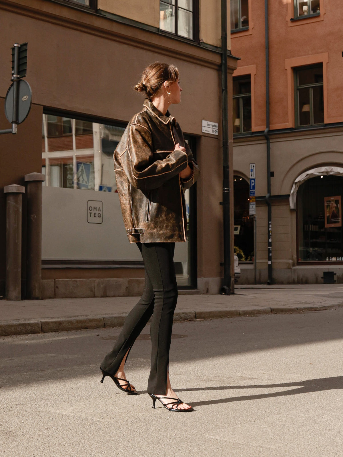 A woman walks along a city street wearing a brown patterned leather jacket, black flared pants, and black strappy high-heeled sandals.