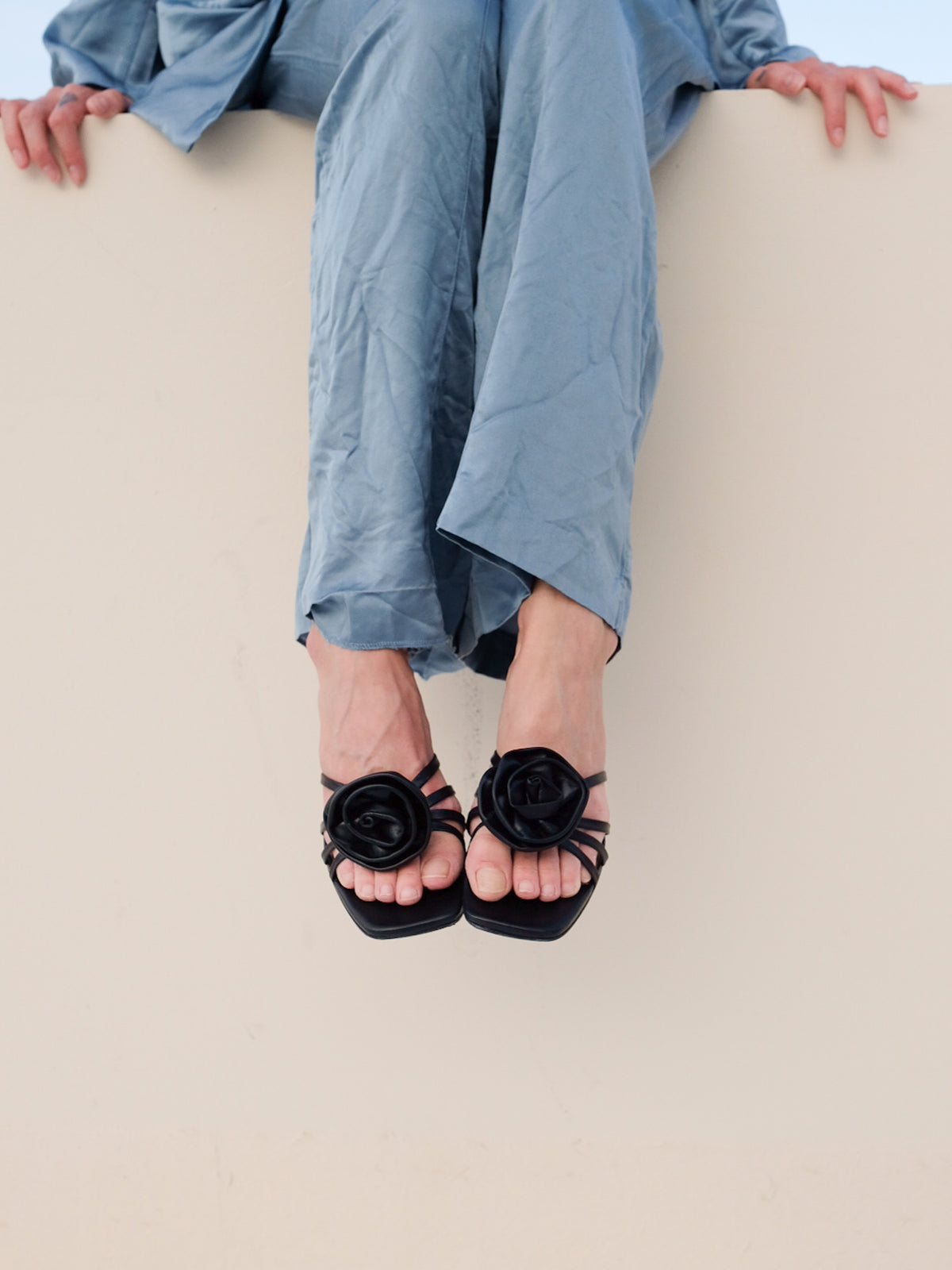 A person wearing black strappy sandals with large black fabric roses on the toes, paired with loose light blue pants, sitting against a plain beige wall.