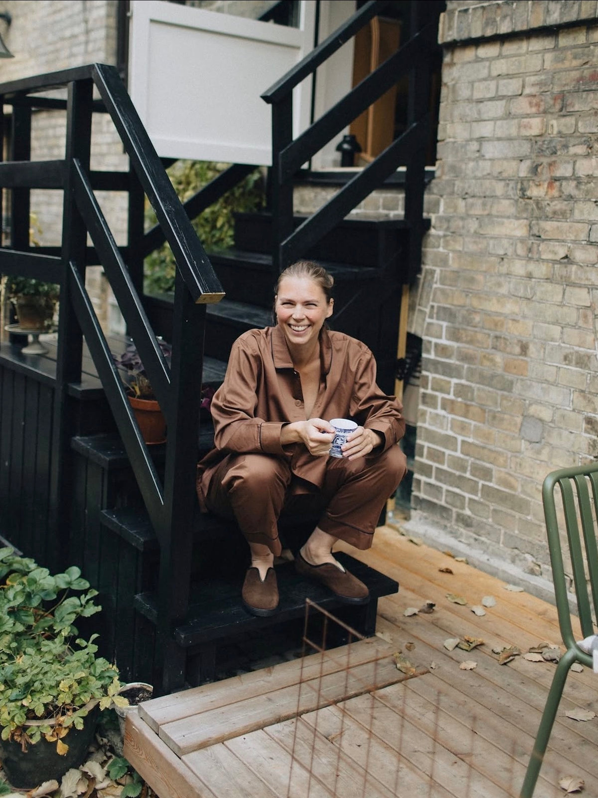 A smiling woman crouches on black outdoor stairs holding a mug, wearing a matching brown lounge set. The setting is a brick house with a wooden deck and plants.