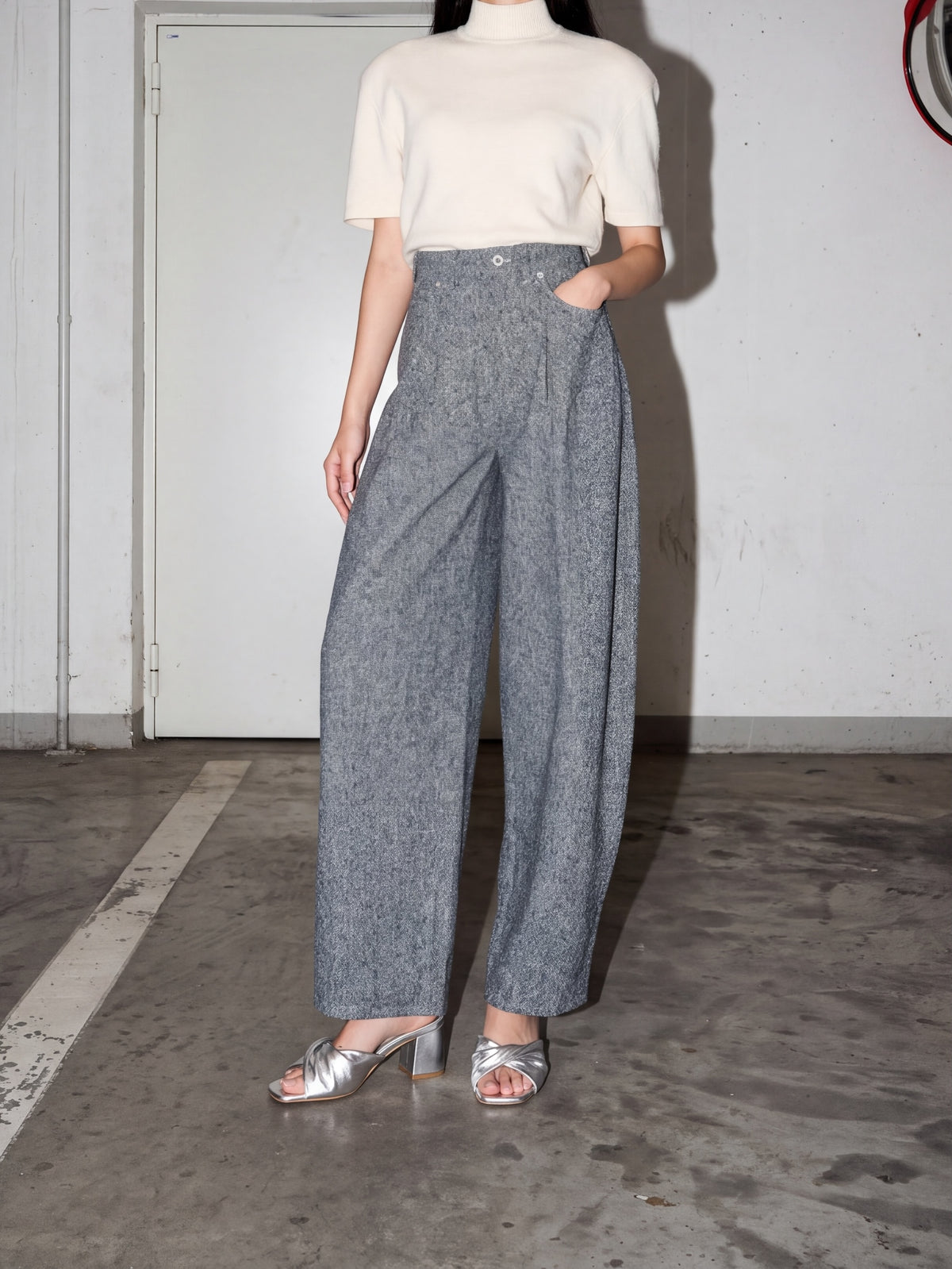 A model wearing a cream short-sleeve mock-neck top and high-waisted, wide-leg gray textured trousers, paired with metallic silver heeled sandals, standing in an indoor concrete space.