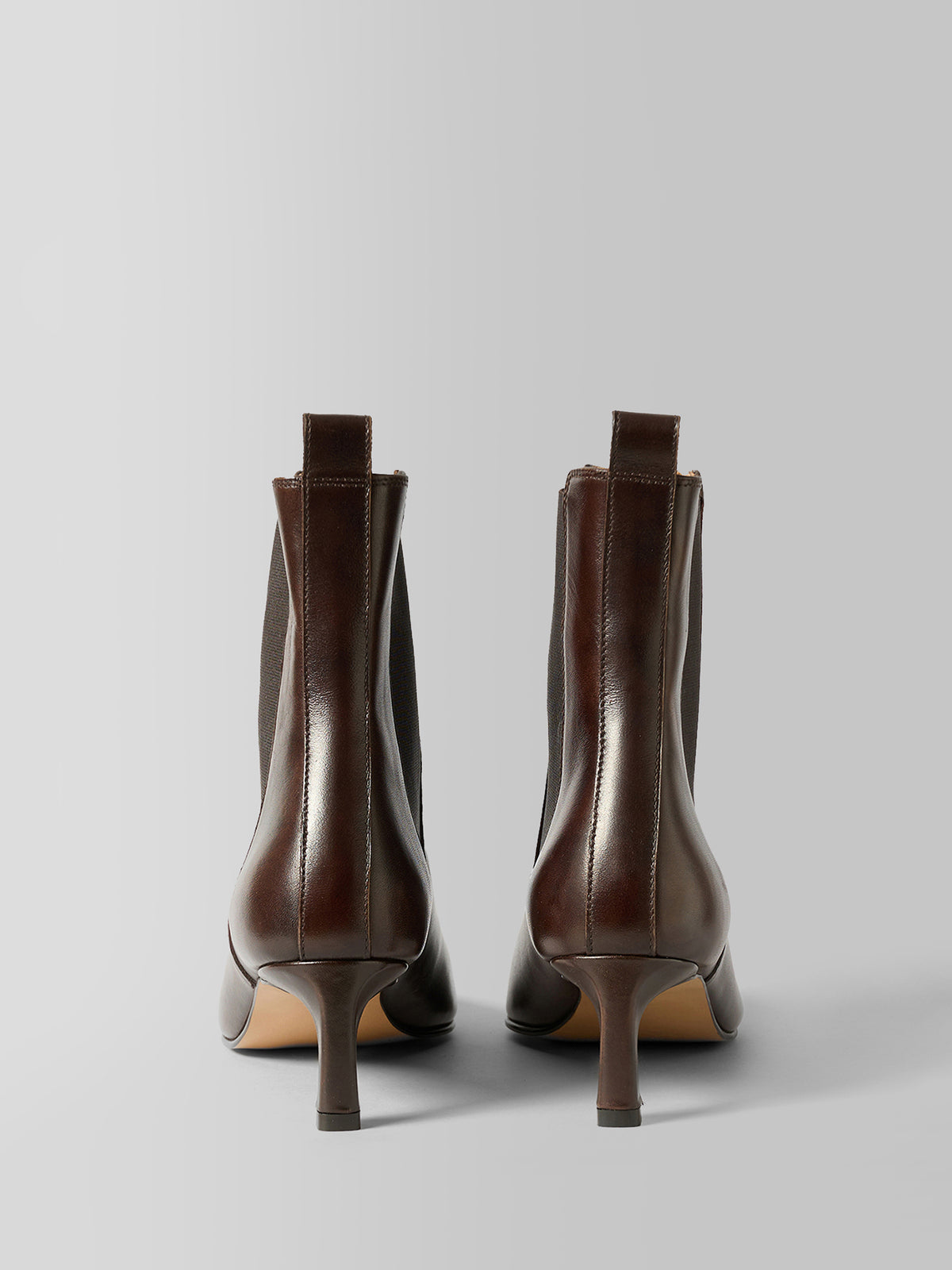 A pair of dark brown leather ankle boots with kitten heels, shown from the back on a light gray studio background.