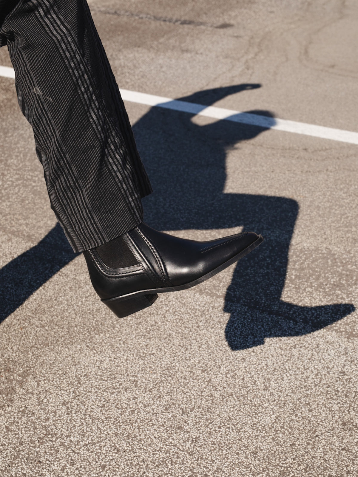 A close-up outdoor shot of a person's leg wearing a black leather pointed-toe boot with a low block heel, standing on asphalt. The person's shadow is cast prominently on the ground.