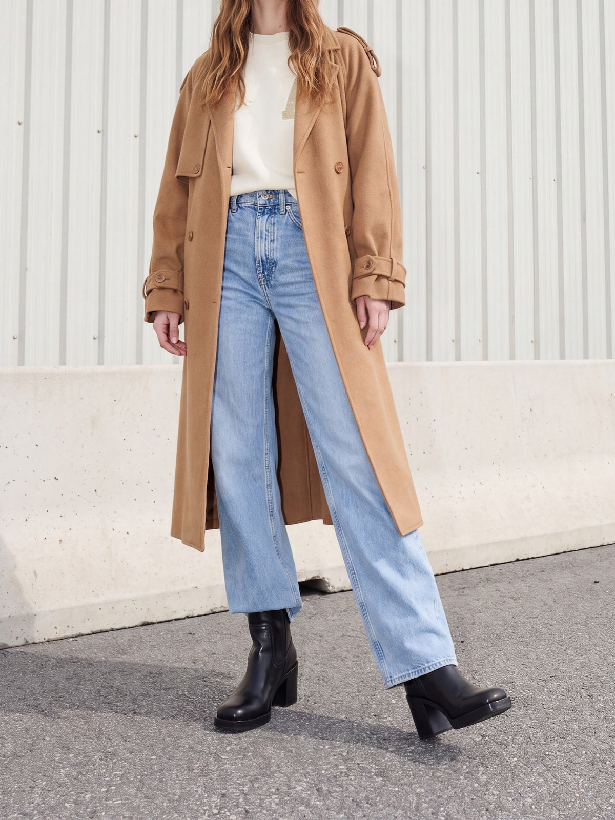 A model wearing a long tan trench coat over a cream top, light blue wide-leg jeans, and black ankle boots, standing outdoors against a corrugated metal wall.