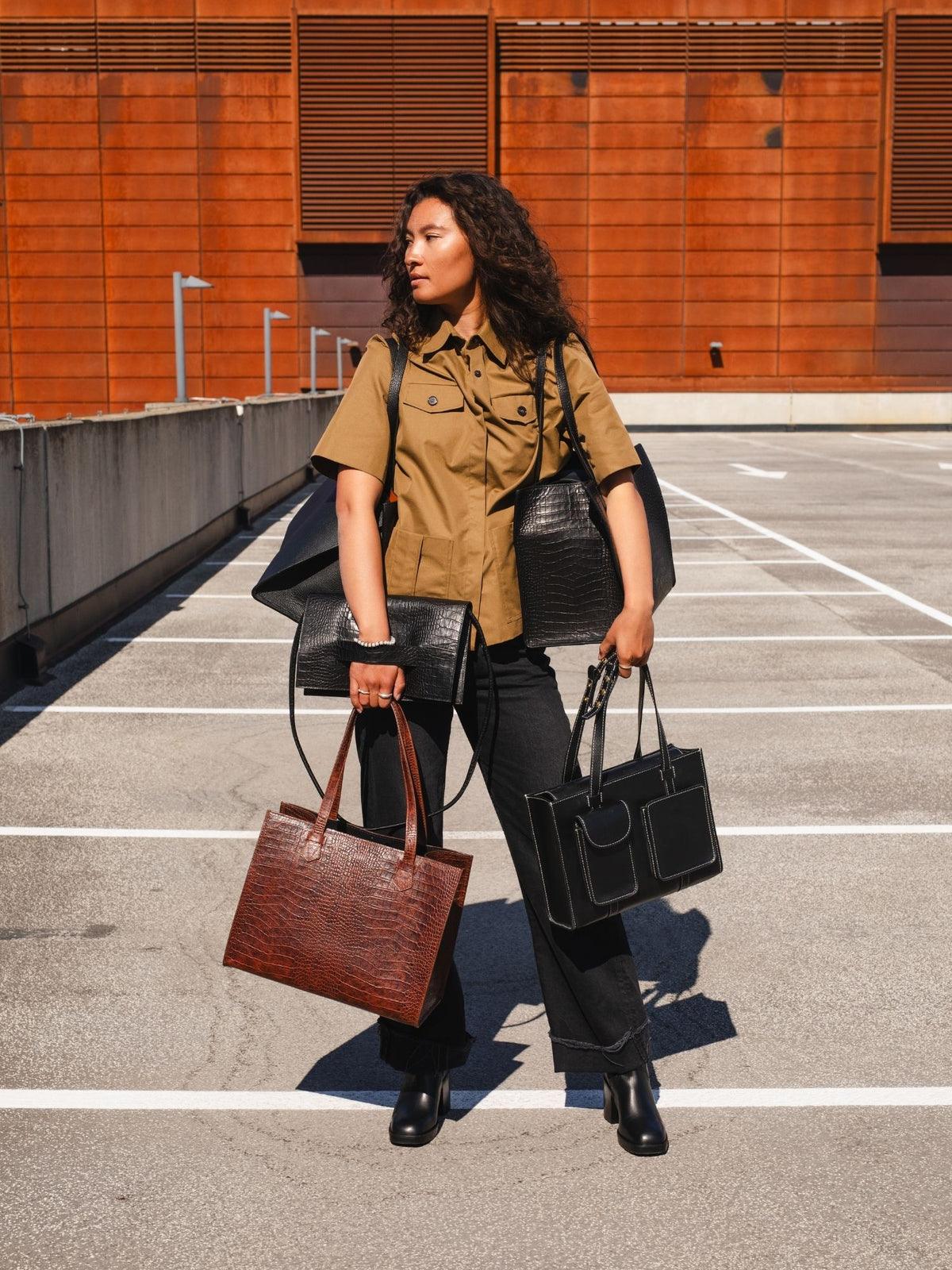 A woman stands in a sunny outdoor parking lot holding multiple handbags. She is wearing a tan short-sleeve button-up shirt, black wide-leg pants, and black boots.