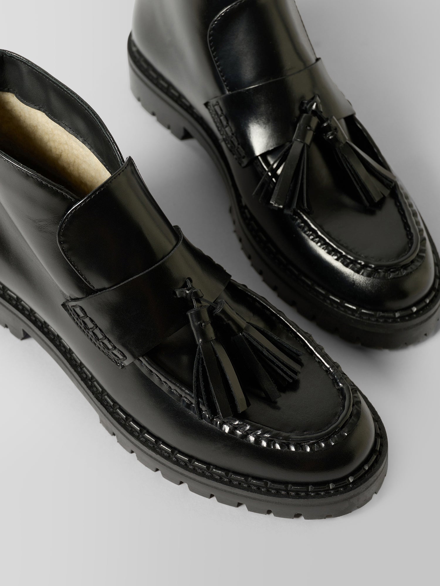 A pair of glossy black leather tassel loafers with thick lug soles, shown on a neutral light gray background.