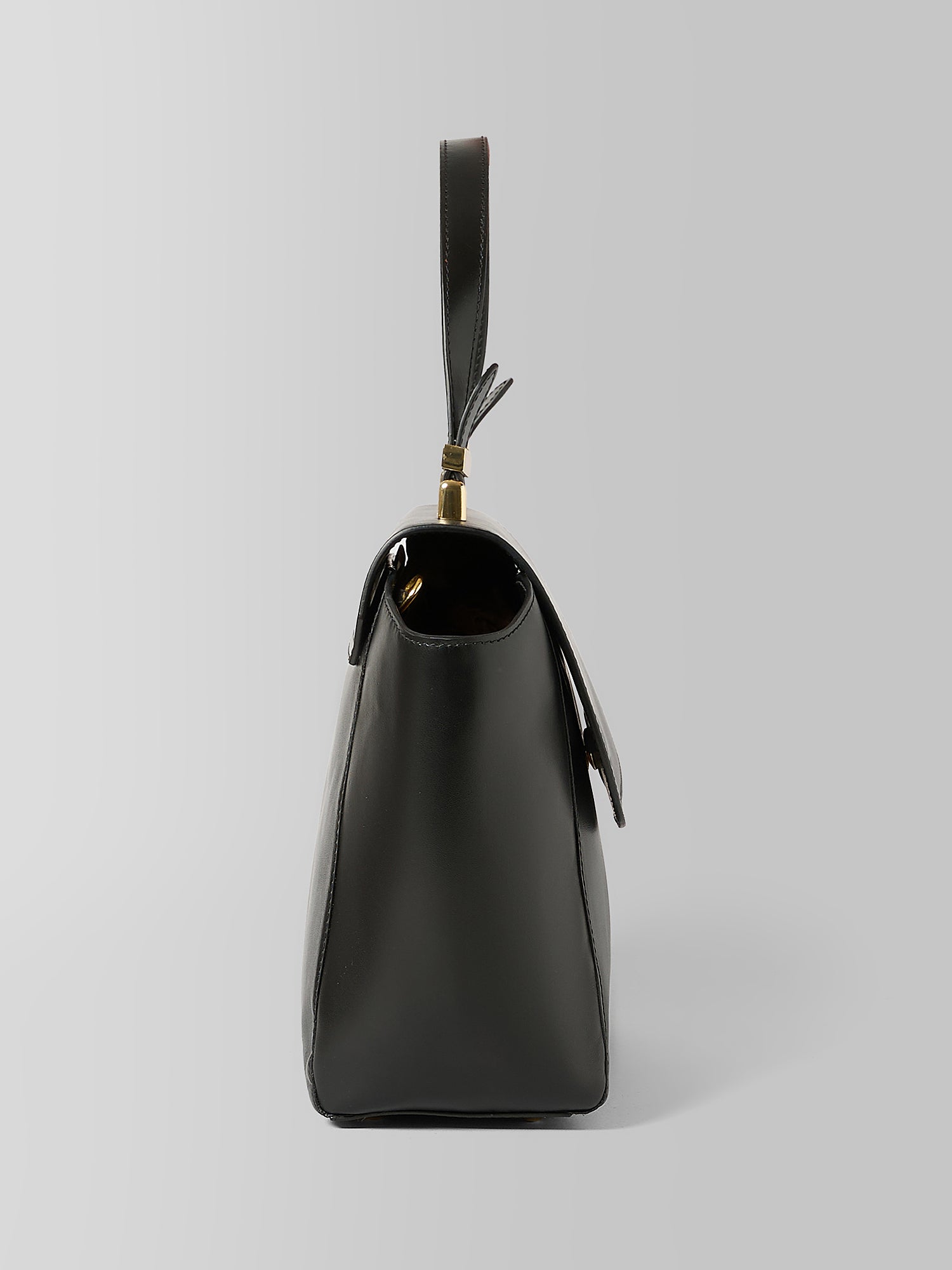 A black leather shoulder bag shown from a side profile view, featuring a top handle with a gold-tone ring and tassel detail, set against a neutral light gray background.