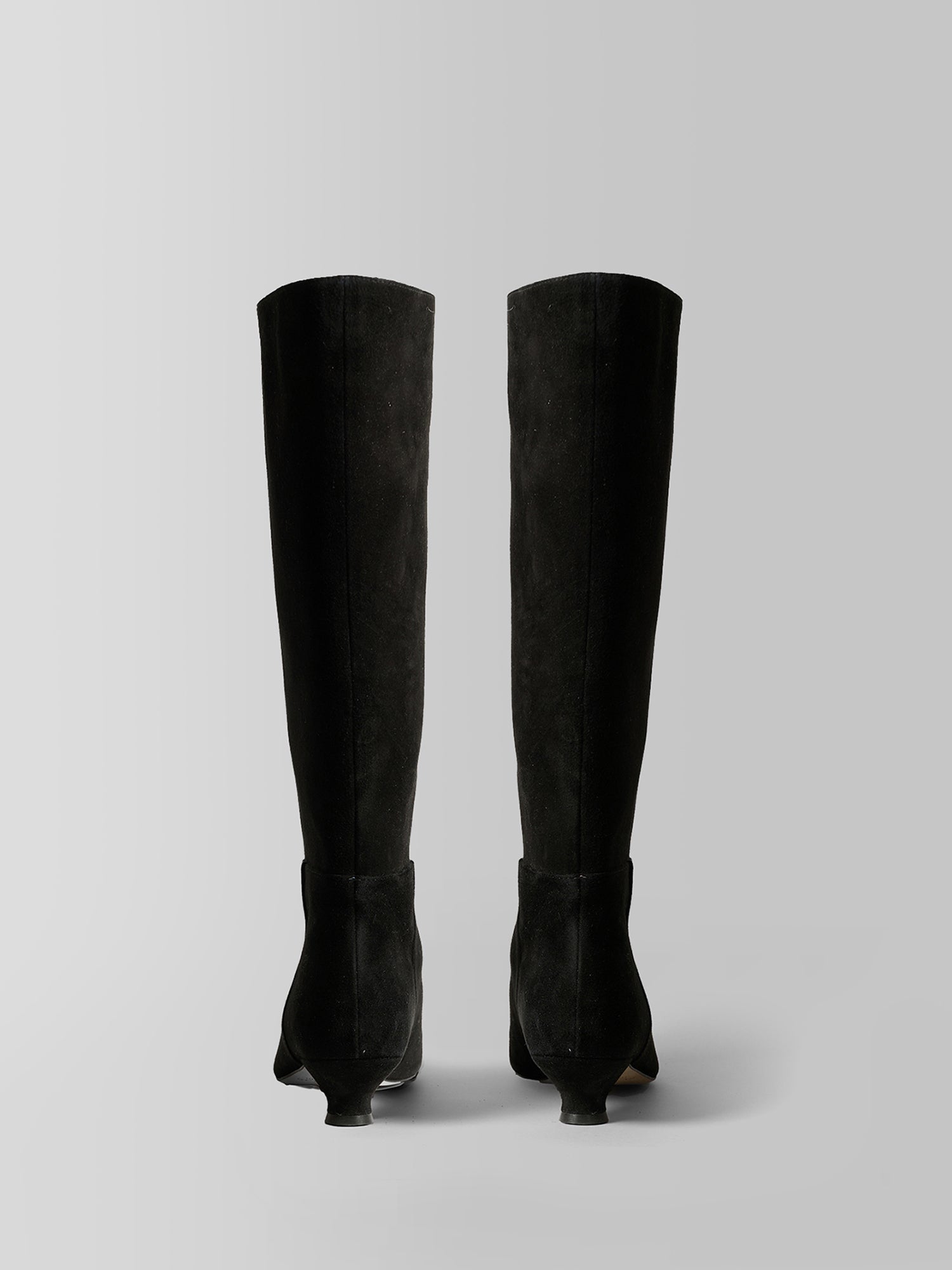 A pair of black suede knee-high boots with pointed kitten heels, shown from the back against a neutral light gray background.