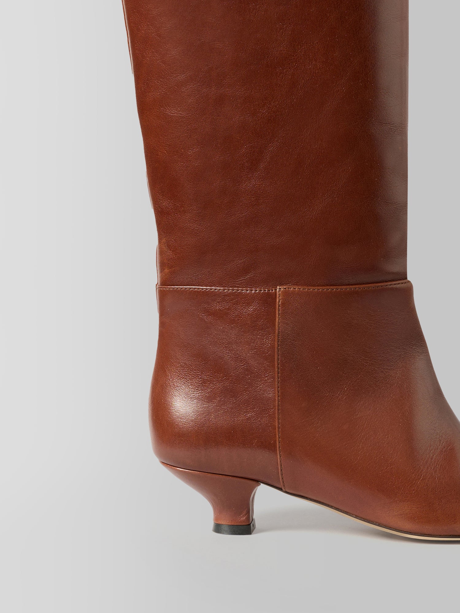 A close-up side view of a brown leather knee-high boot with a low kitten heel, set against a neutral light gray background.