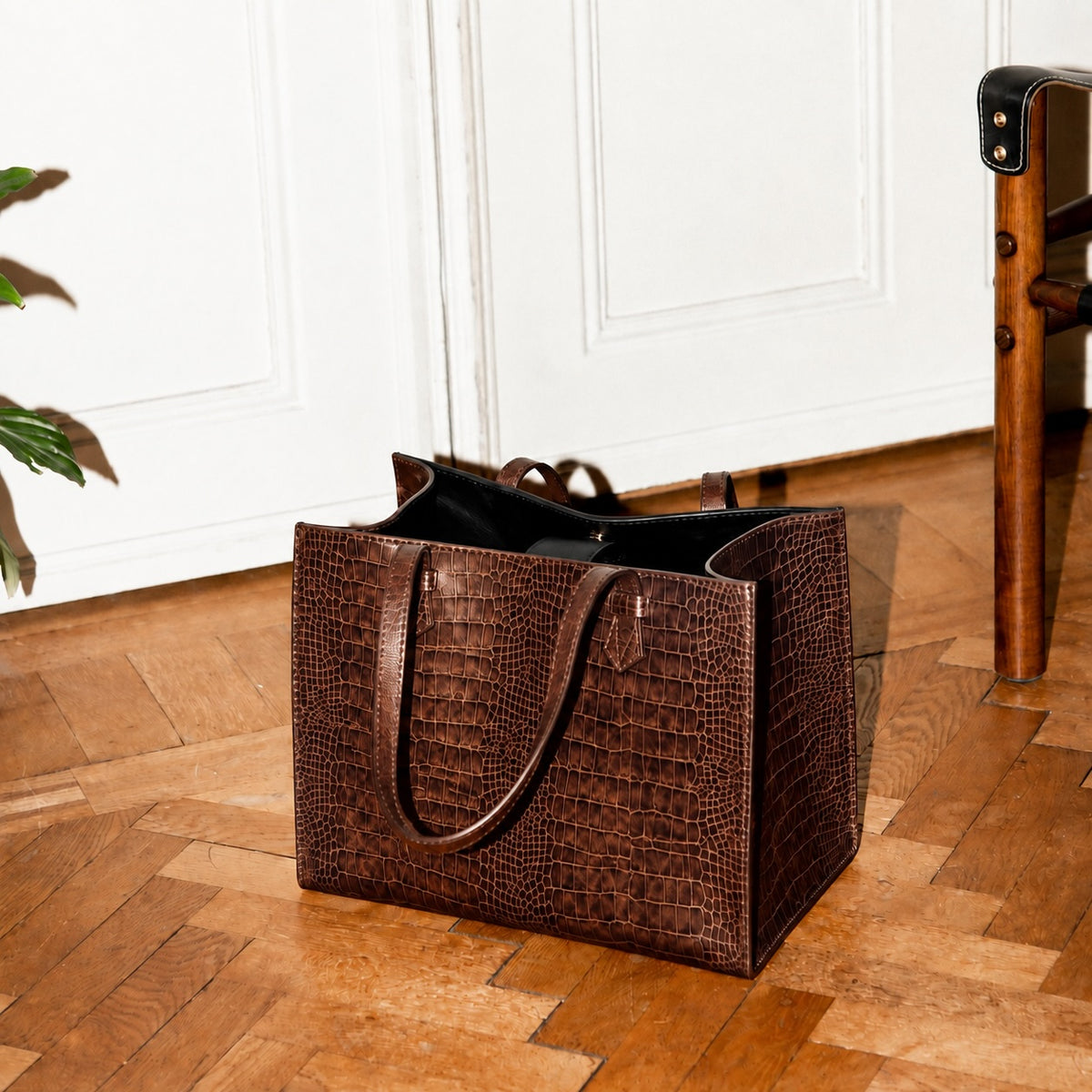 A dark brown crocodile-embossed leather tote bag with matching shoulder straps, resting on a polished wooden parquet floor in front of white paneled doors.