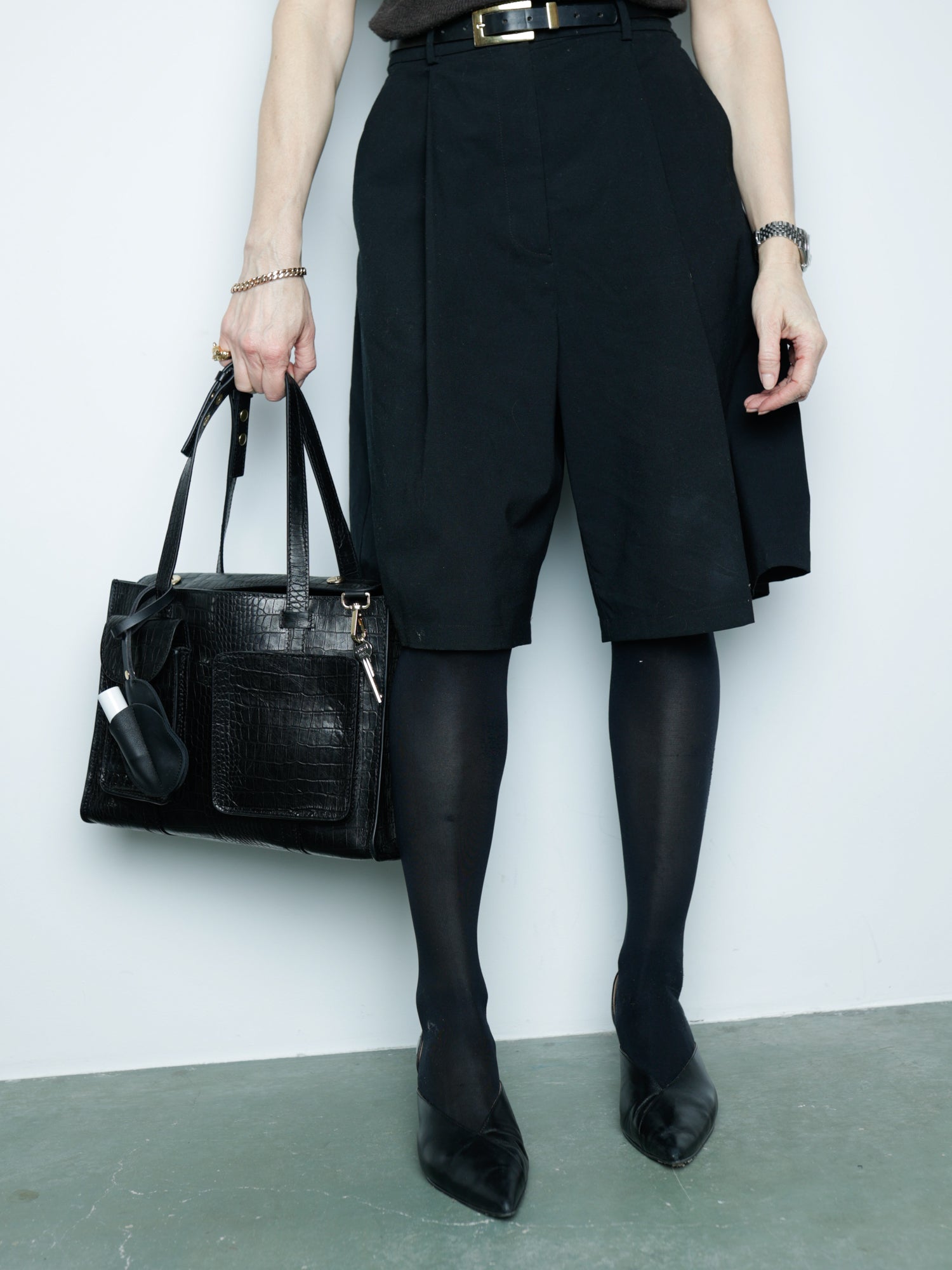 Person wearing black shorts, tights, and shoes holding a black handbag against a white wall.