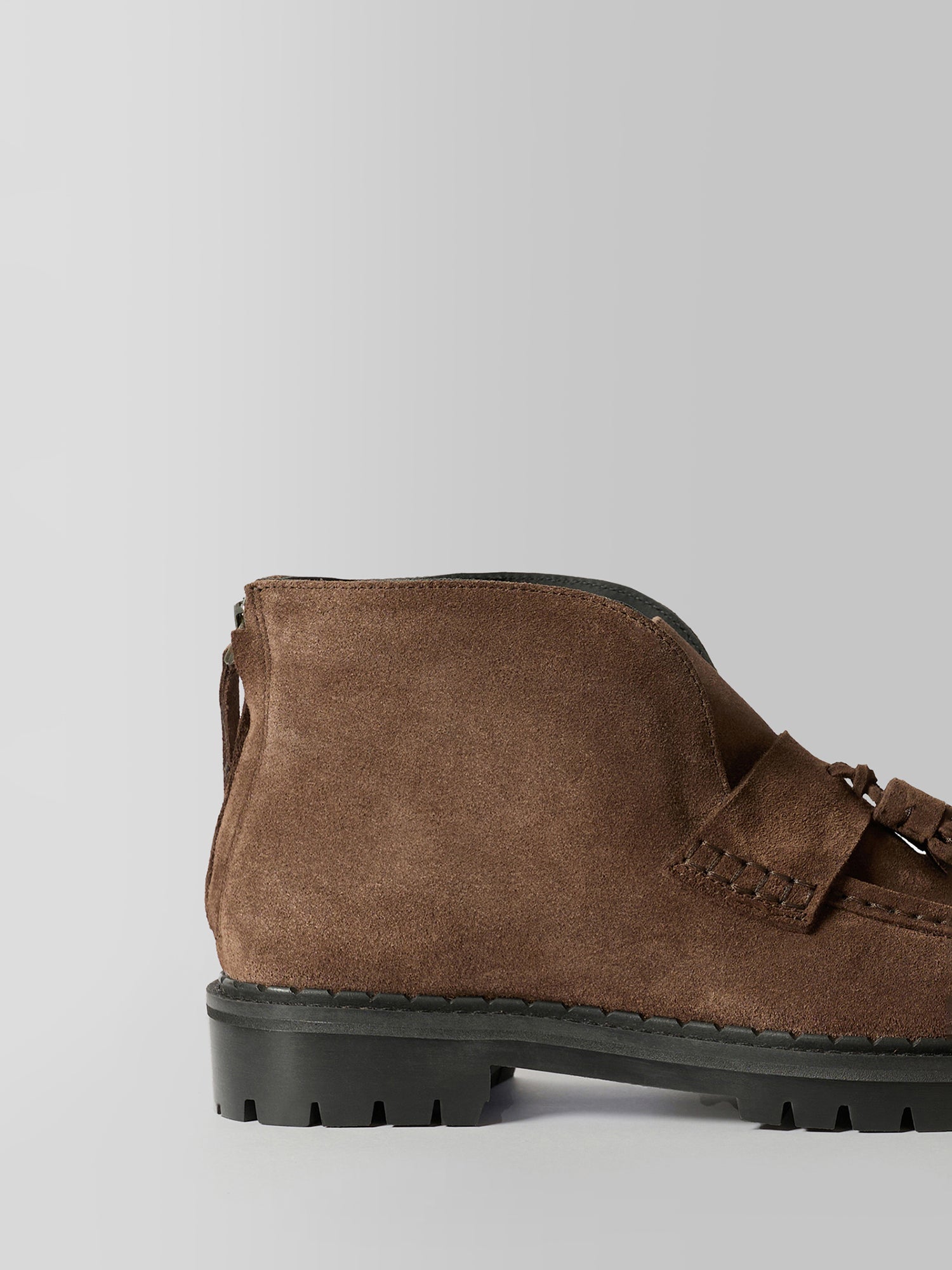 A close-up side view of a brown suede ankle boot with a black lug sole and a decorative strap detail on the side, set against a plain light gray background.