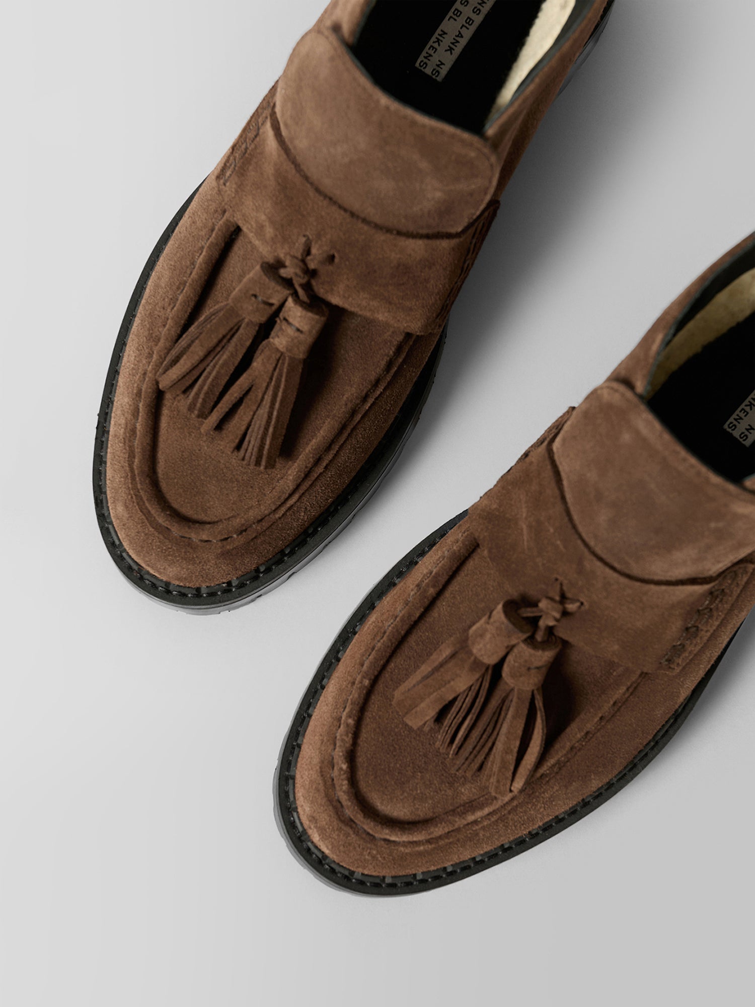 A pair of brown suede penny loafers with tassel details, shown from a top-down angle on a light gray background.