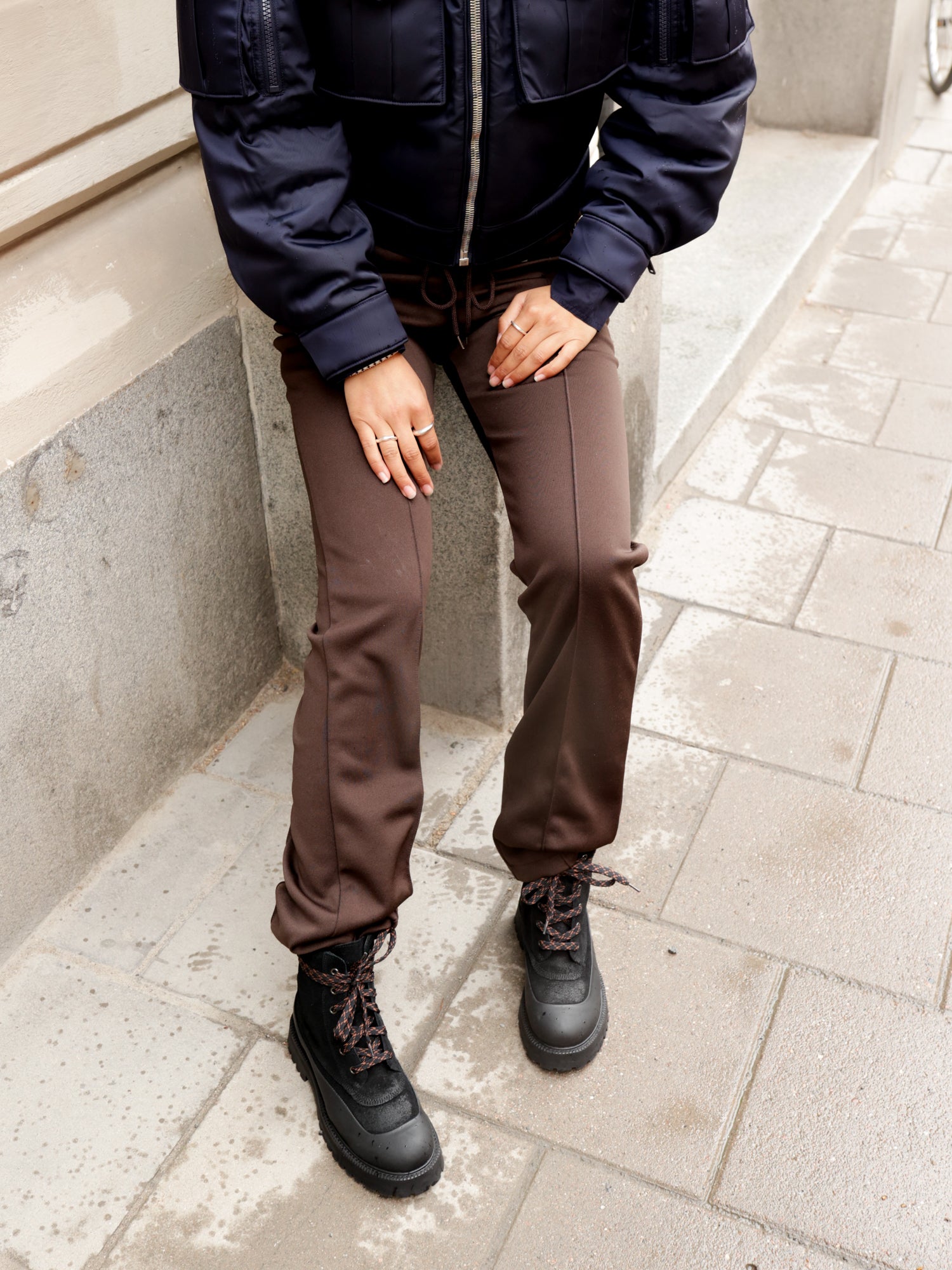 Person wearing a dark jacket, brown pants, and black boots on a sidewalk.