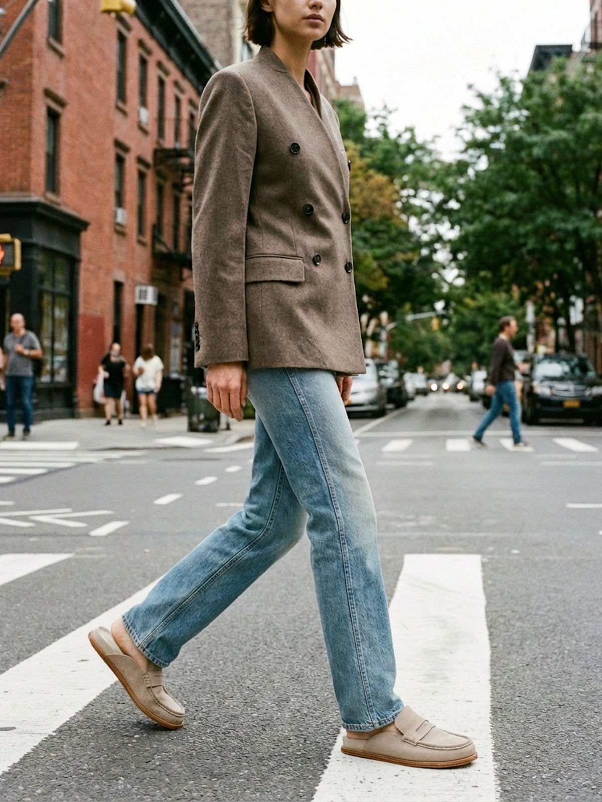 Person walking on a city street wearing a brown blazer, blue jeans, and beige shoes.