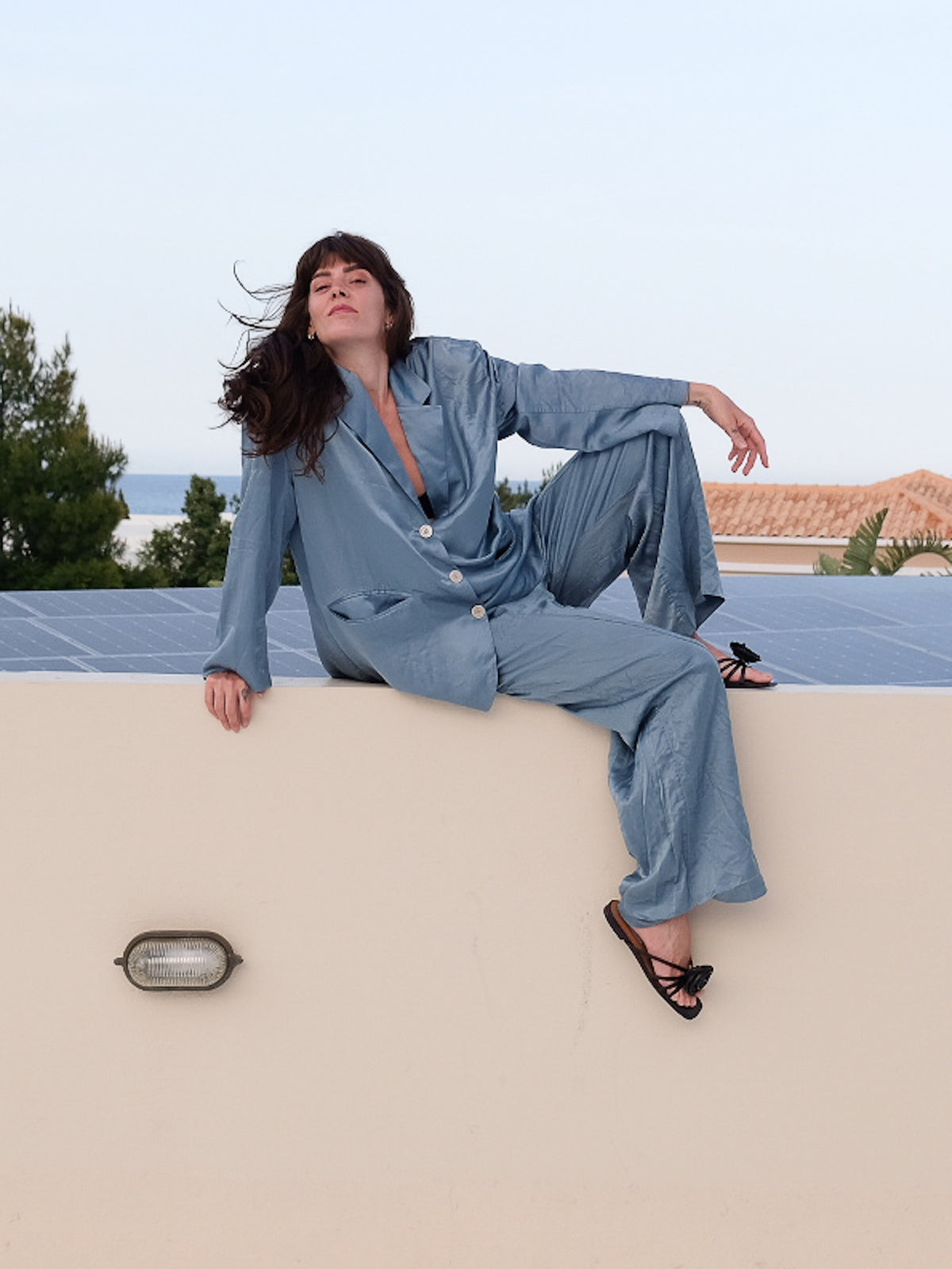 A woman wearing a light blue satin pajama set with a button-down shirt and matching pants, sitting on a beige wall outdoors with solar panels and the sea in the background.