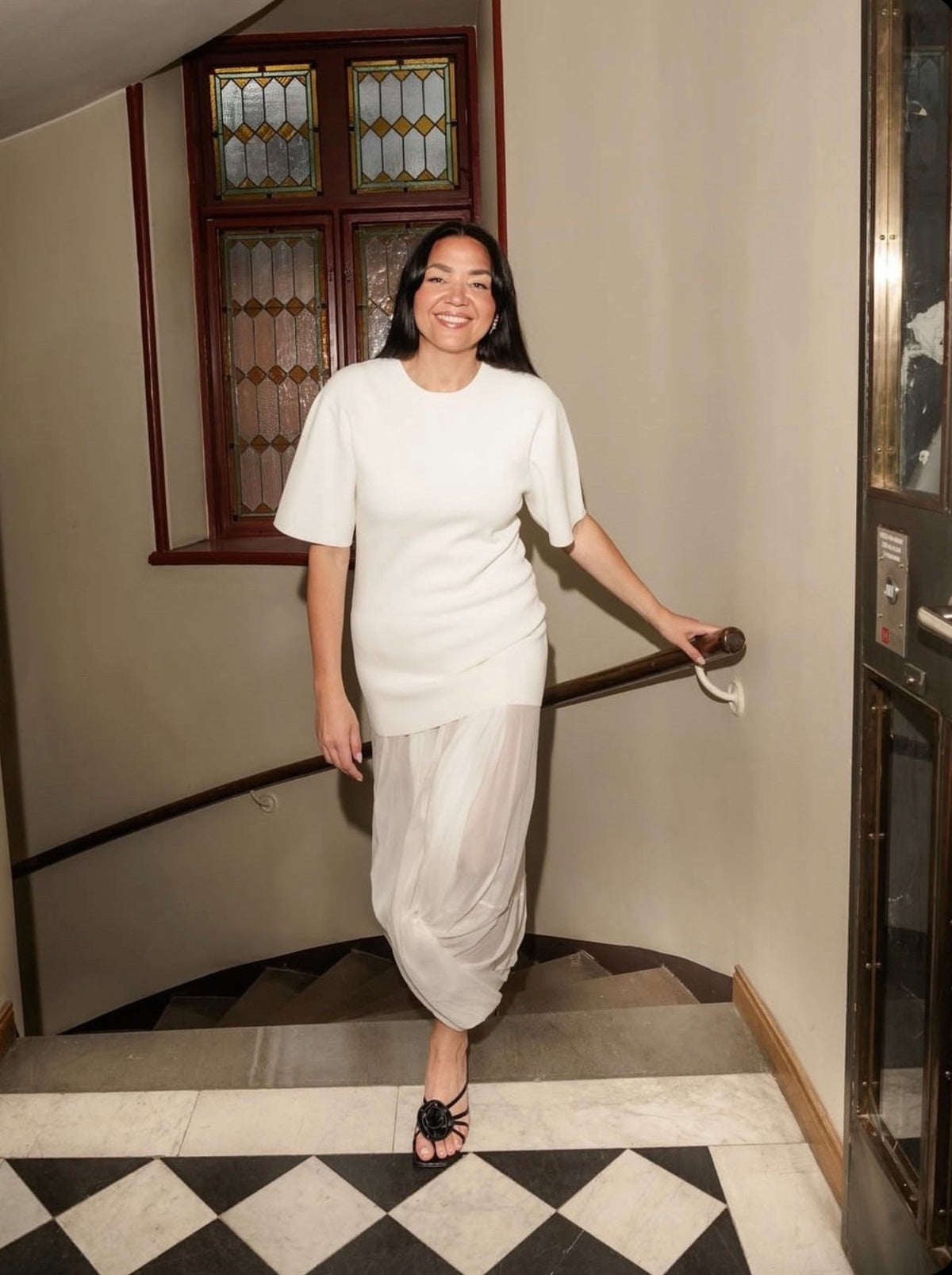 A woman wearing a white short-sleeve top and matching draped white skirt stands on an indoor staircase with a black-and-white checkered floor, holding a handrail.