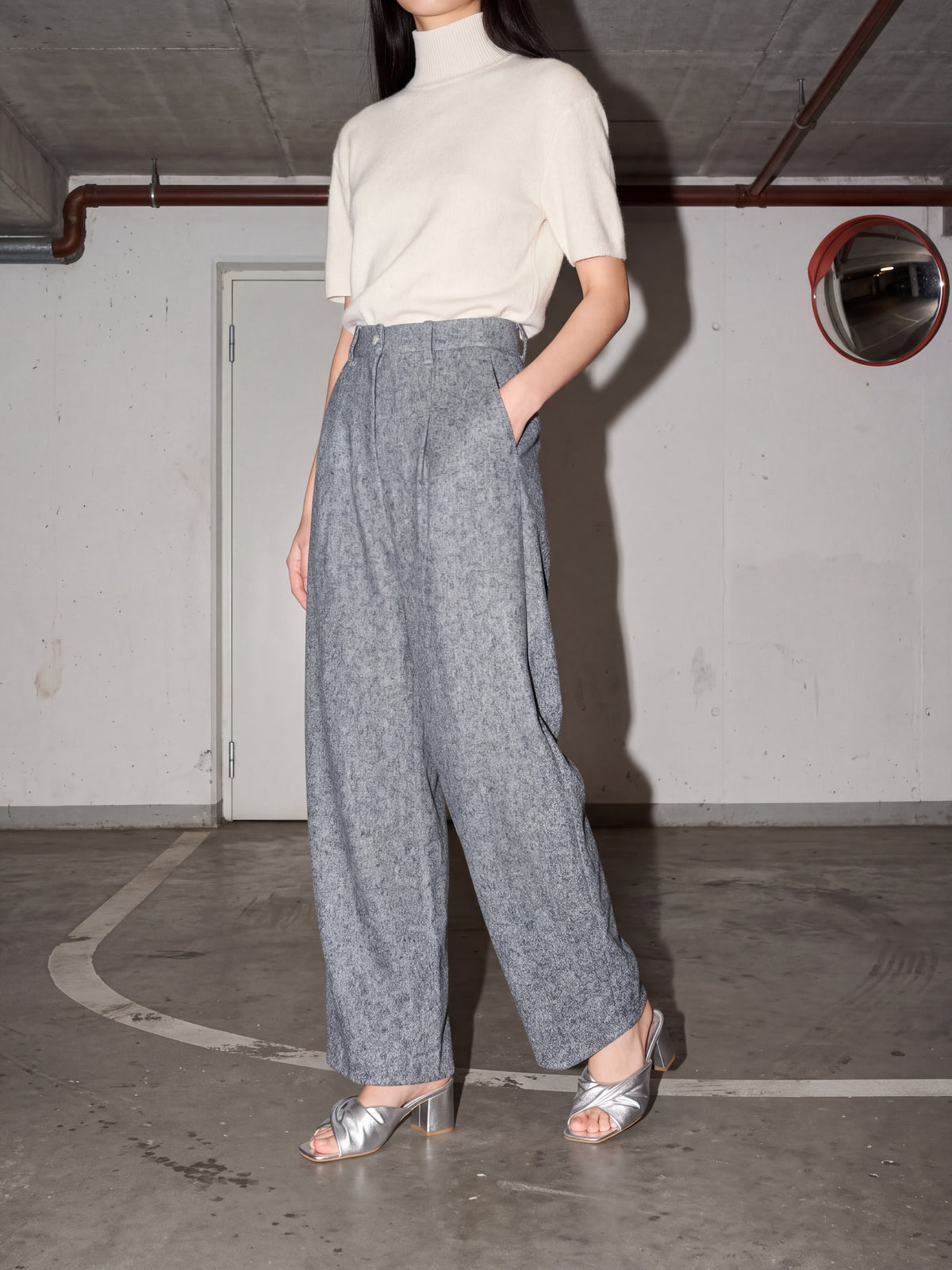A model wearing a cream short-sleeve mock neck top and high-waisted, wide-leg gray textured trousers, paired with silver heeled sandals, standing in a concrete parking garage.