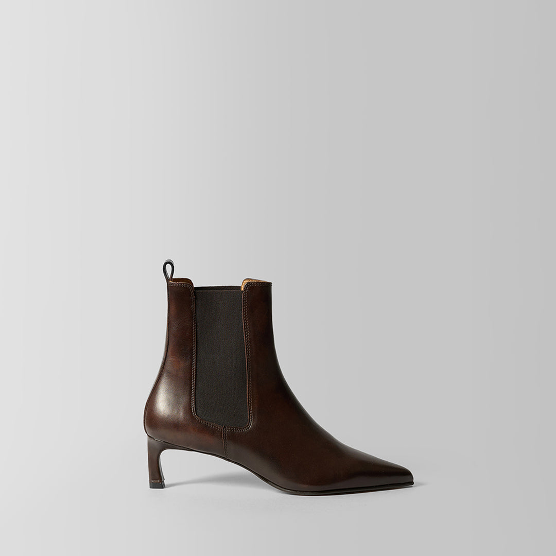 A dark brown leather ankle boot with a pointed toe and a dark gray elastic side panel, shown from a side profile on a light gray background.