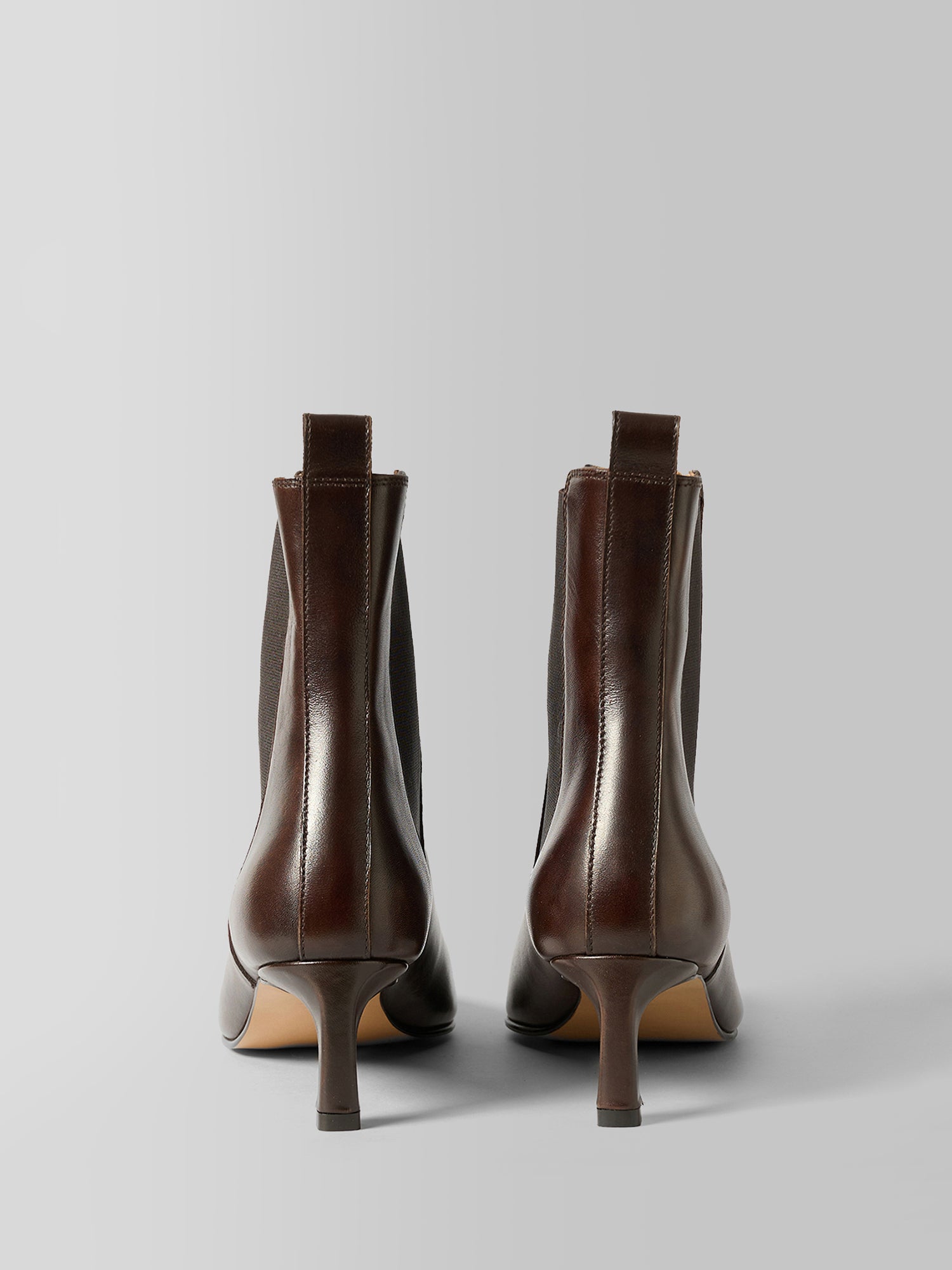 A pair of dark brown leather ankle boots with kitten heels, shown from the back on a light gray studio background.
