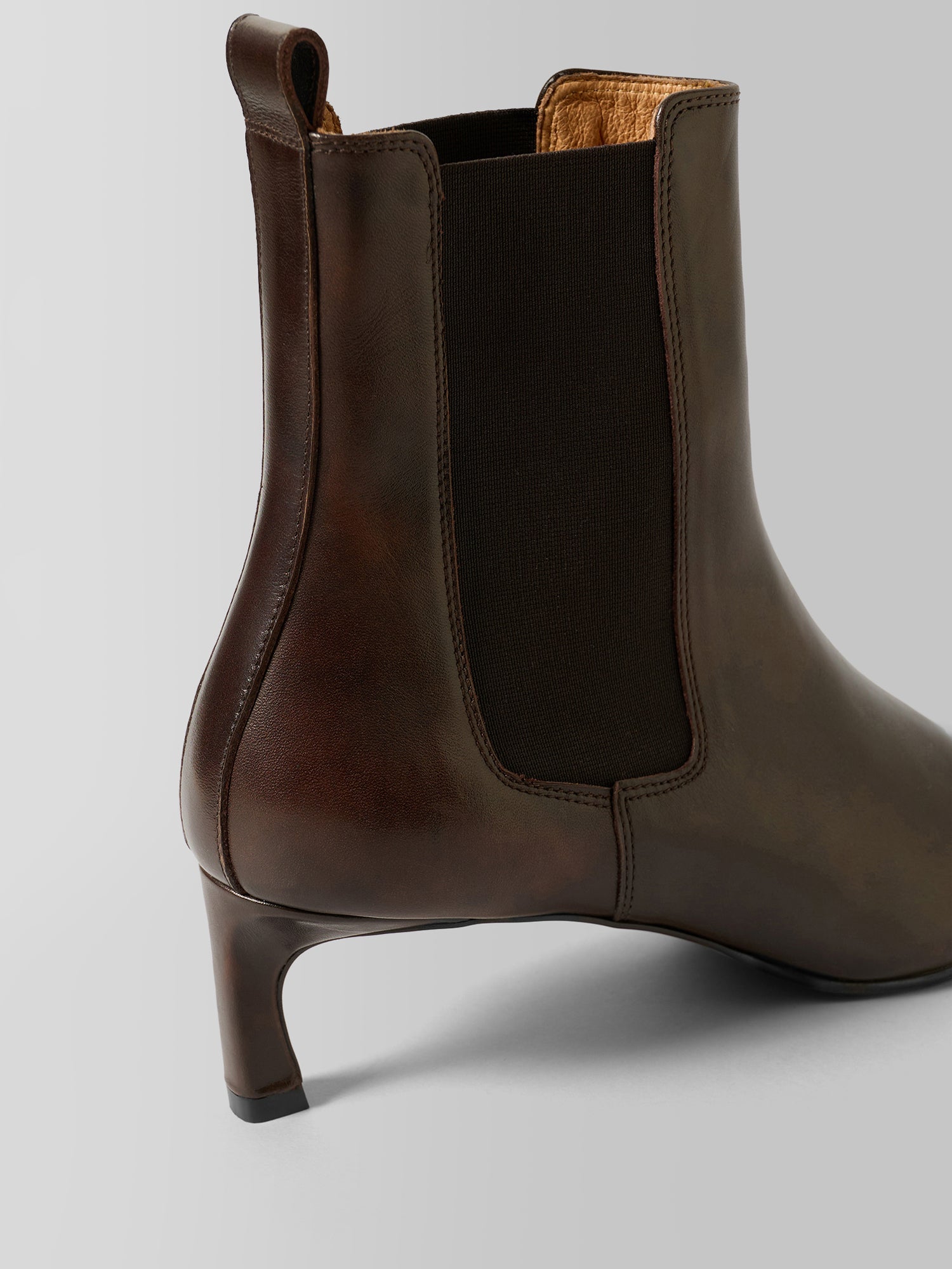 A dark brown leather Chelsea boot with a dark brown elastic side panel and a low kitten heel, shown from a three-quarters rear angle on a light gray background.