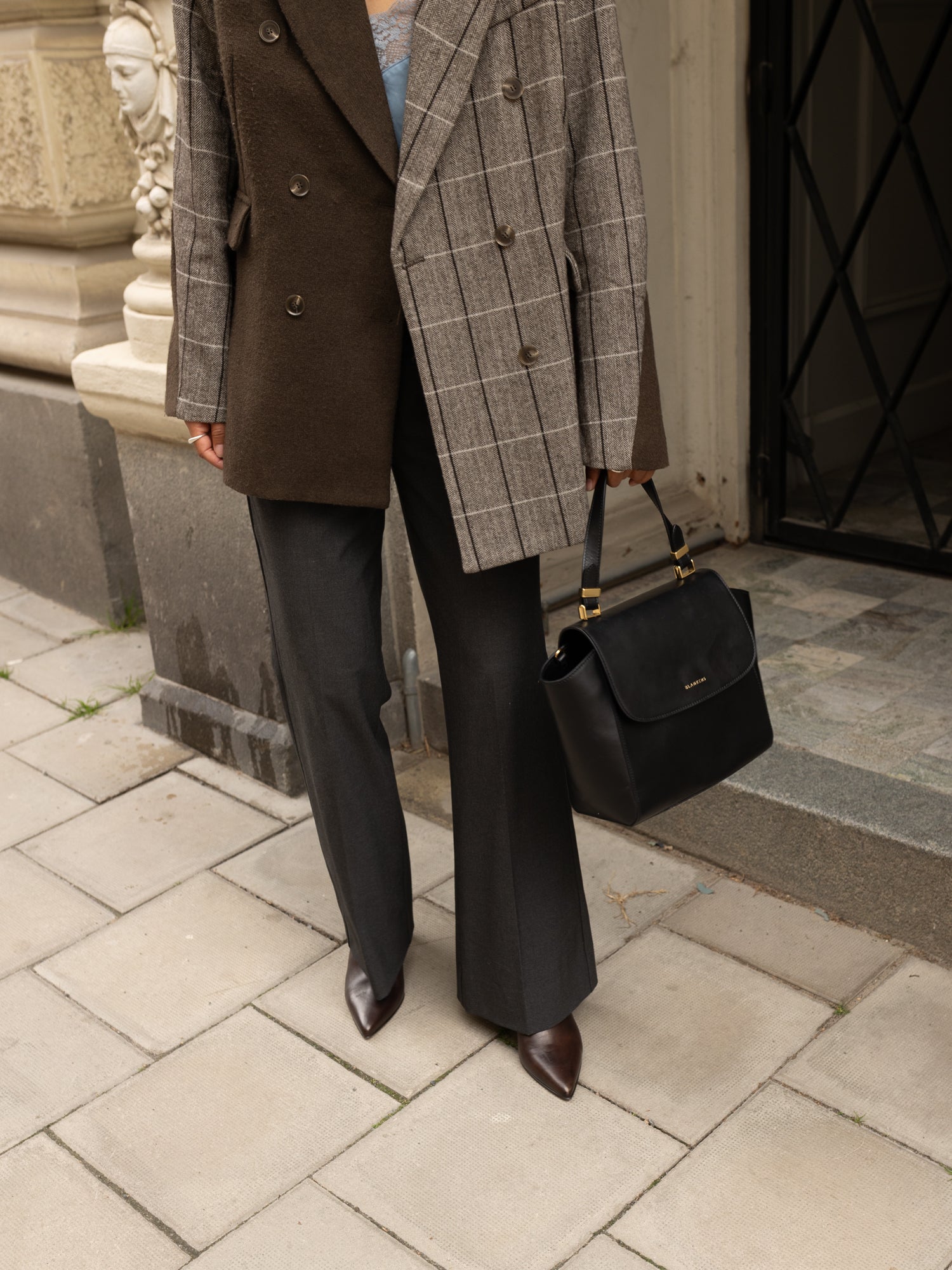 Person wearing a brown and gray checkered blazer with a black handbag on a stone pavement.