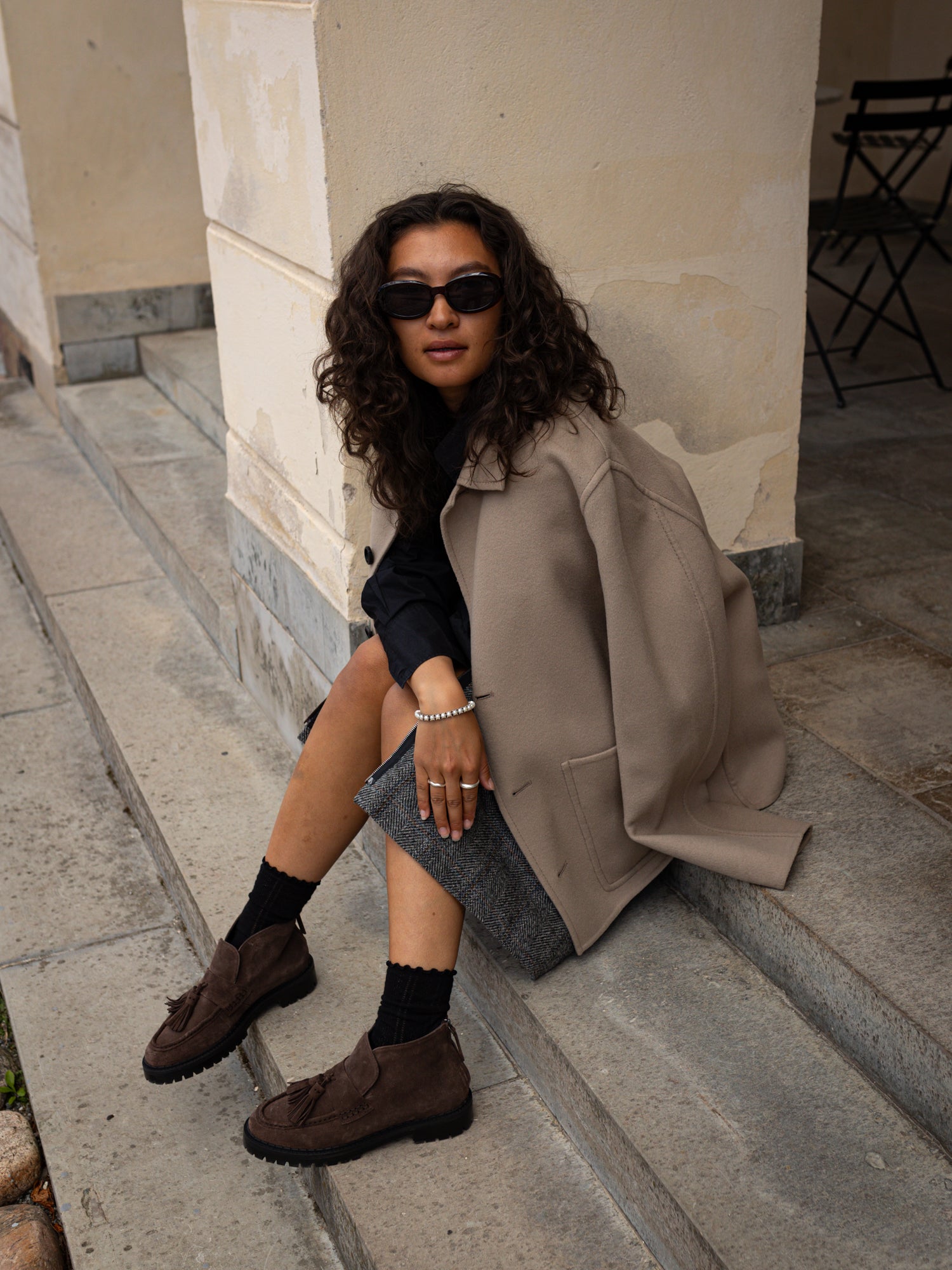 A woman sits on stone steps wearing a large taupe blazer, dark top, gray skirt, brown suede loafers, and black socks in an outdoor urban setting.