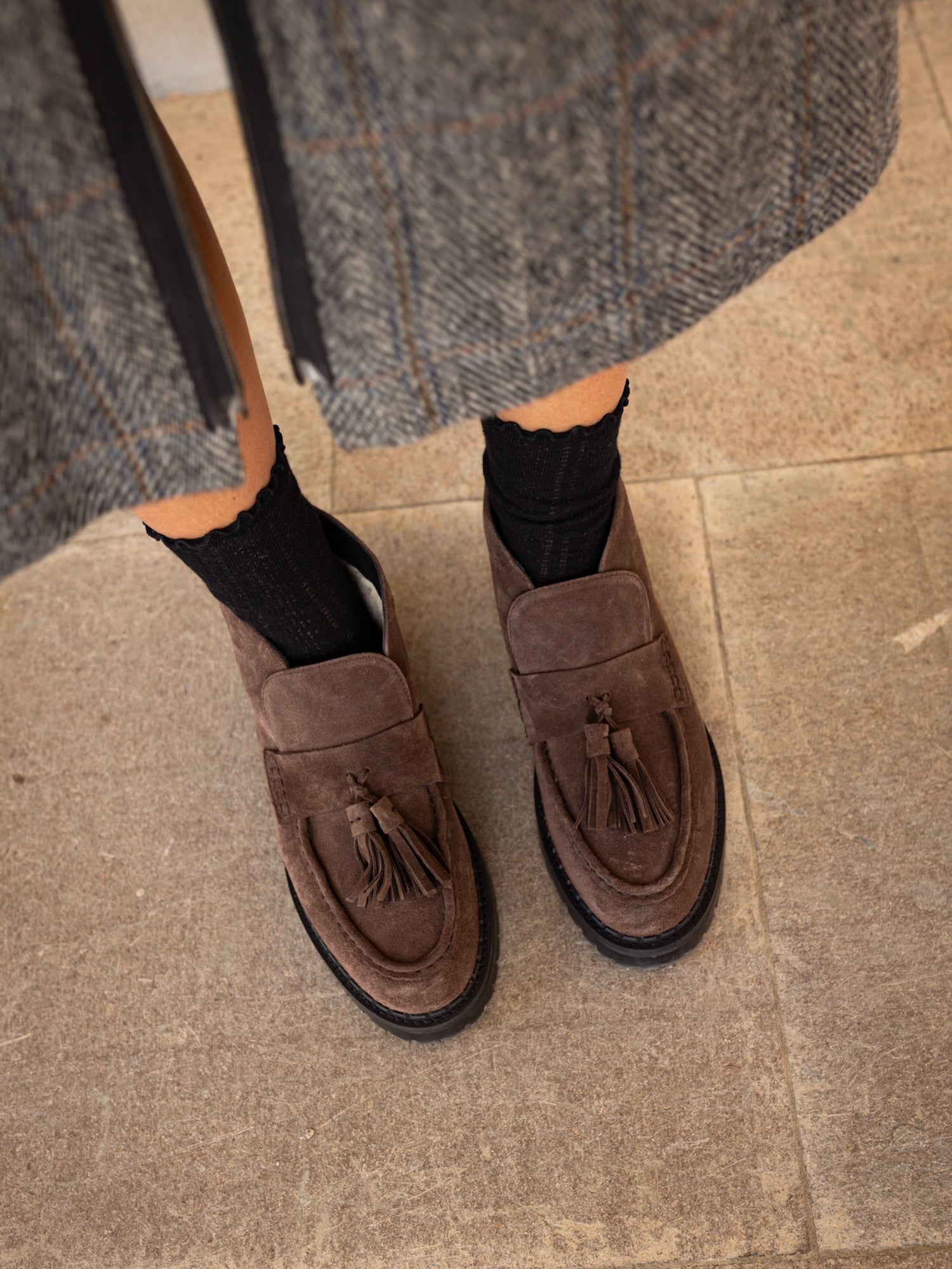 A pair of brown suede tassel loafers worn with black crew socks, shown from a top-down angle on a beige tiled floor with a gray herringbone skirt visible at the top.