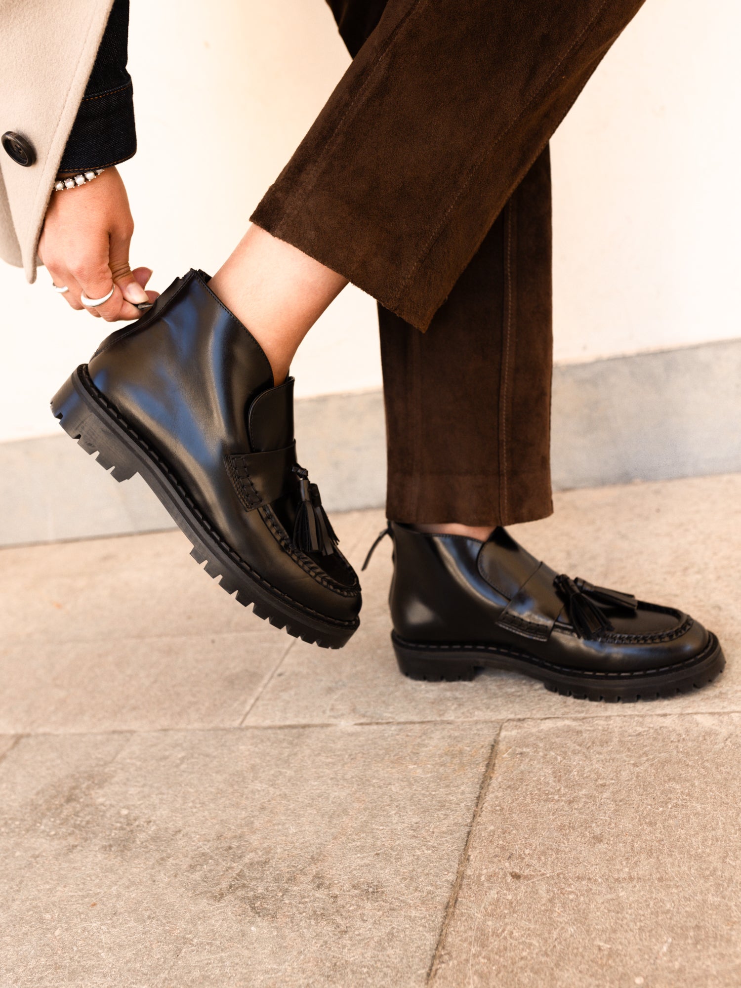 A person wearing black leather tassel loafers with chunky lug soles, paired with brown trousers and a light-colored coat, standing on a gray stone floor.