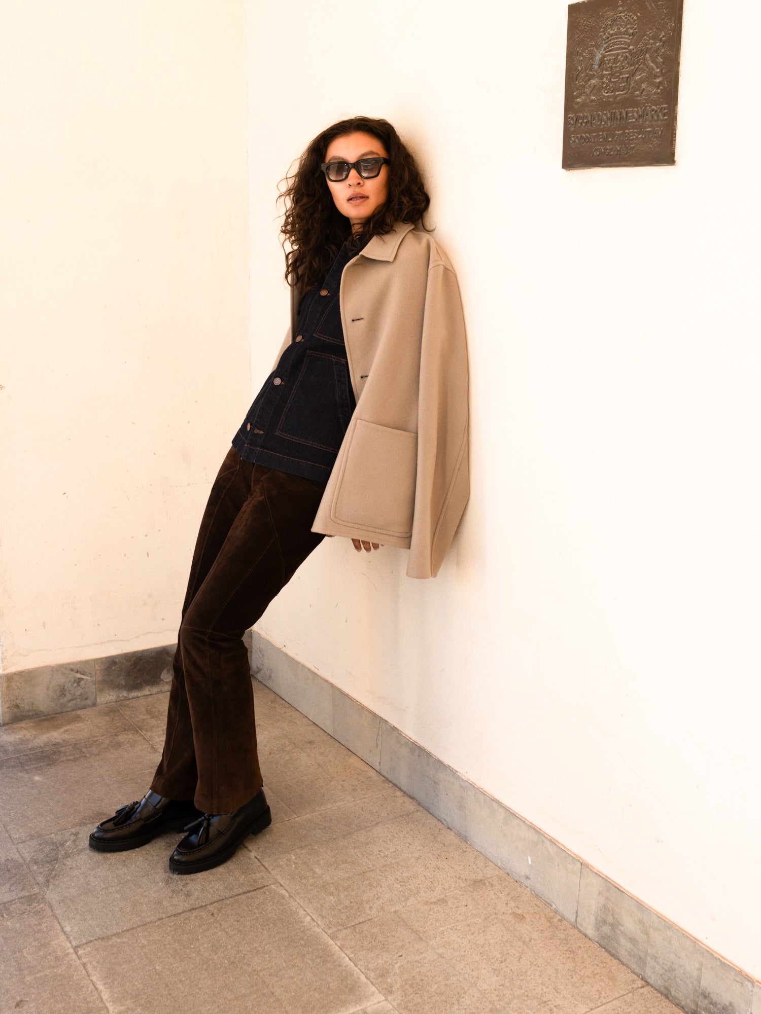 A woman wearing a beige trench coat over a dark denim top and brown trousers, leaning against a cream-colored wall in a minimalist indoor setting.