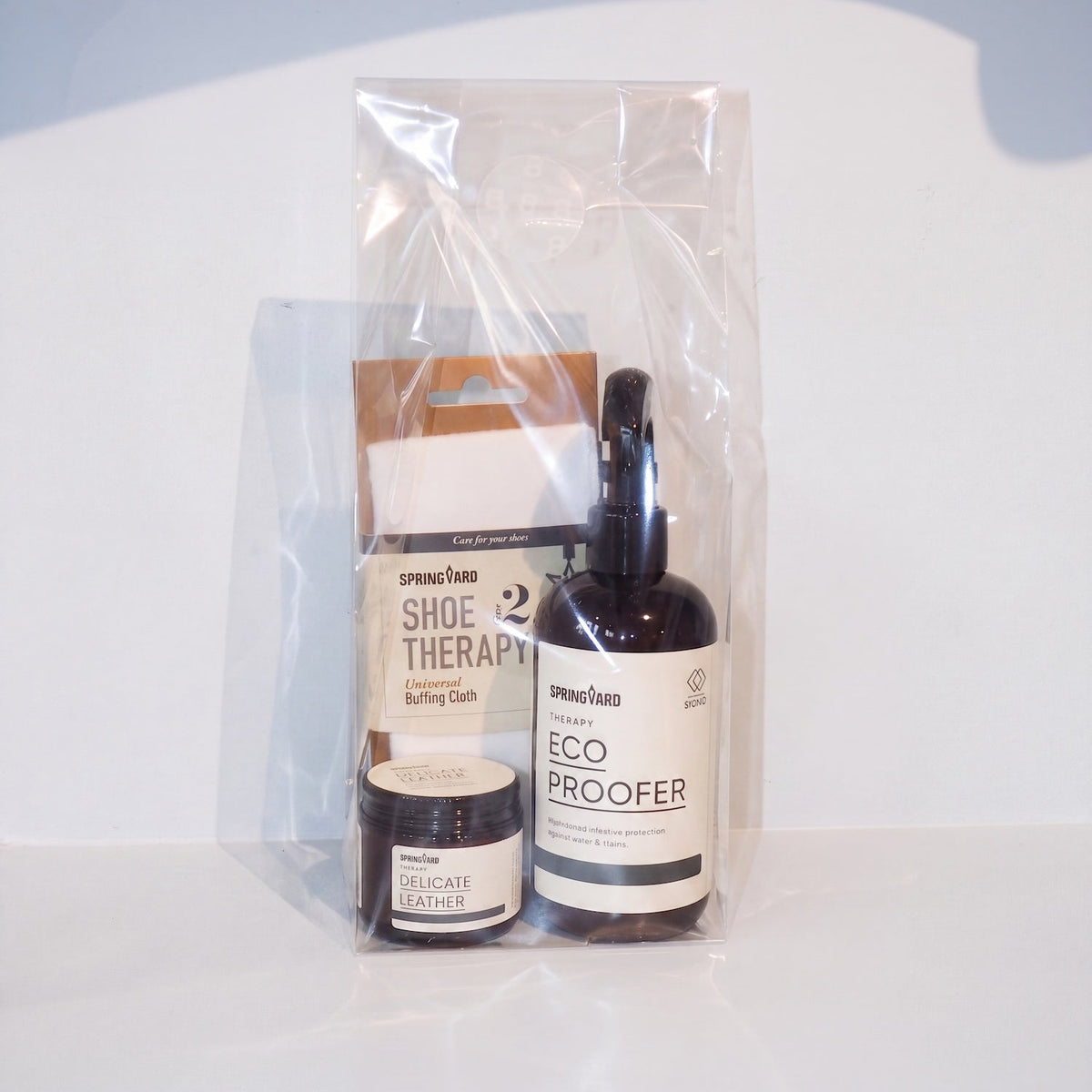 A clear plastic gift bag containing a small jar of leather cleaner, a pump bottle labeled Eco Proofer, and a box labeled Shoe Therapy, on a white surface with a light blue-gray background.