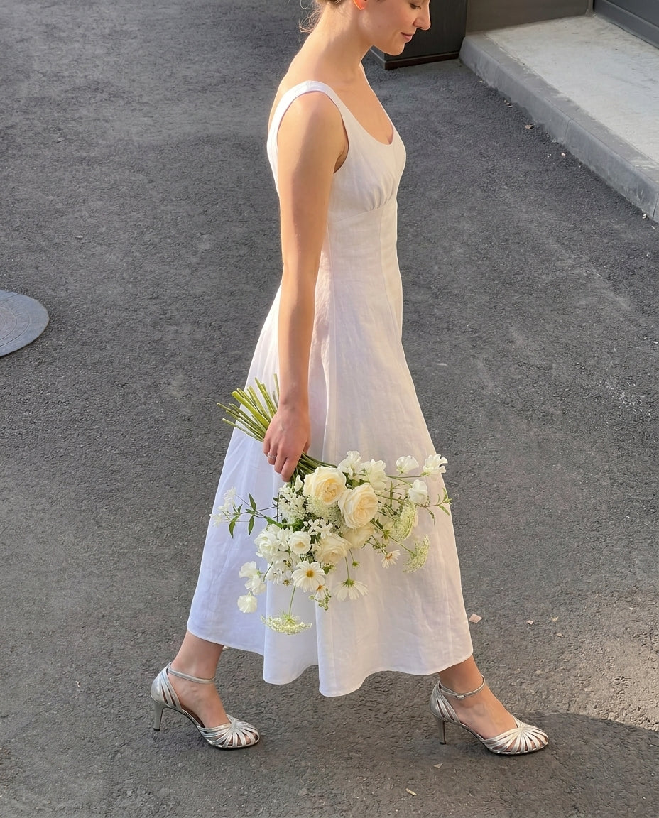 A woman wearing a sleeveless white wedding dress with silver high heels walks outdoors while holding a bouquet of white and cream flowers.