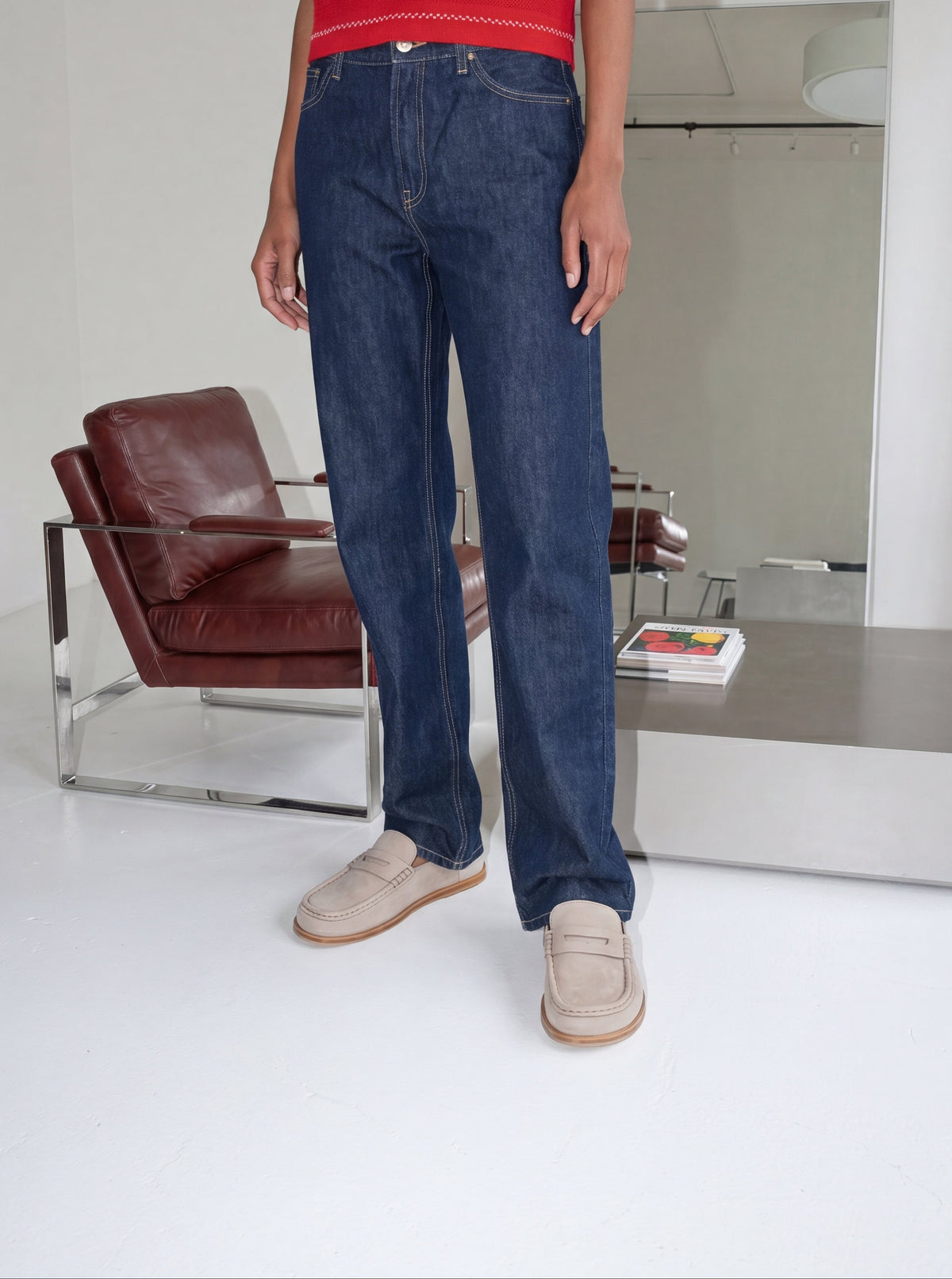 A person wearing dark blue straight-leg denim jeans, a red top, and beige slip-on shoes, standing in a modern minimalist room with a brown leather chair and a mirror.